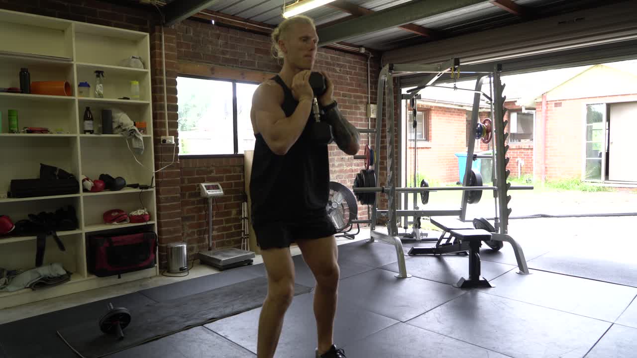 Uomo tatuato nella palestra di casa, ripreso frontalmente mentre esegue goblet squat