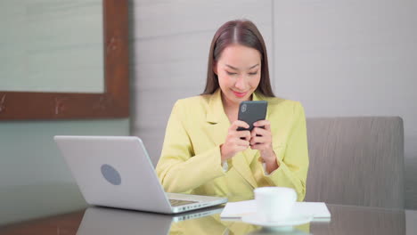 asian businesswoman typing on the screen of mobile phone while sitting at the office desk in front of laptop computer