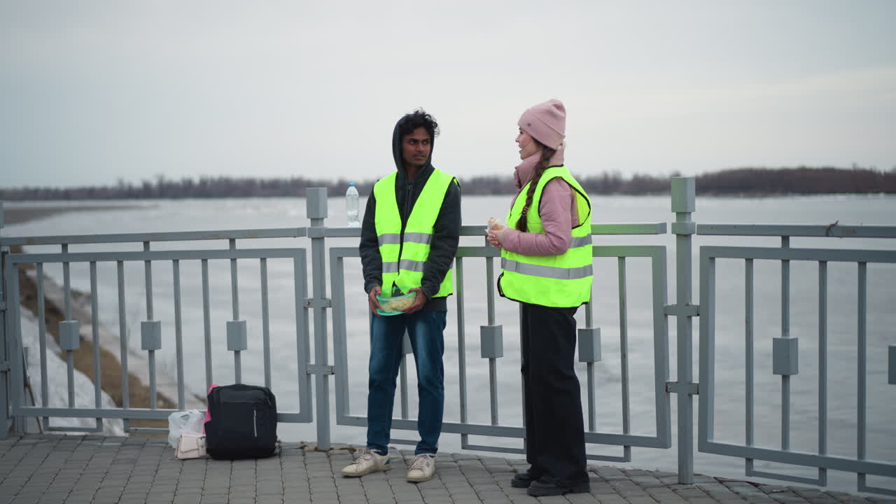 Man in hoodie and neon safety vest holding food container standing with woman in pink jacket and matching vest holding wrap on riverside promenade near metal railing during cold overcast day
