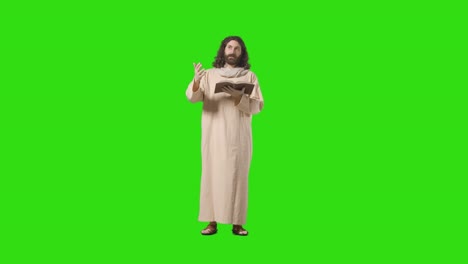 studio shot of man wearing robes and sandals with long hair and beard representing figure of jesus christ preaching from bible on green screen