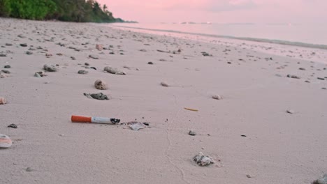 Brennende-Rauchende-Rauchende-Zigarettenkippe,-Die-Auf-Sand-An-Einem-Ziemlich-Leeren,-Unbewohnten-Strand-Bei-Morgendämmerung-Verschmutzung-Müll-Nahaufnahme-Zeitlupe-Fällt