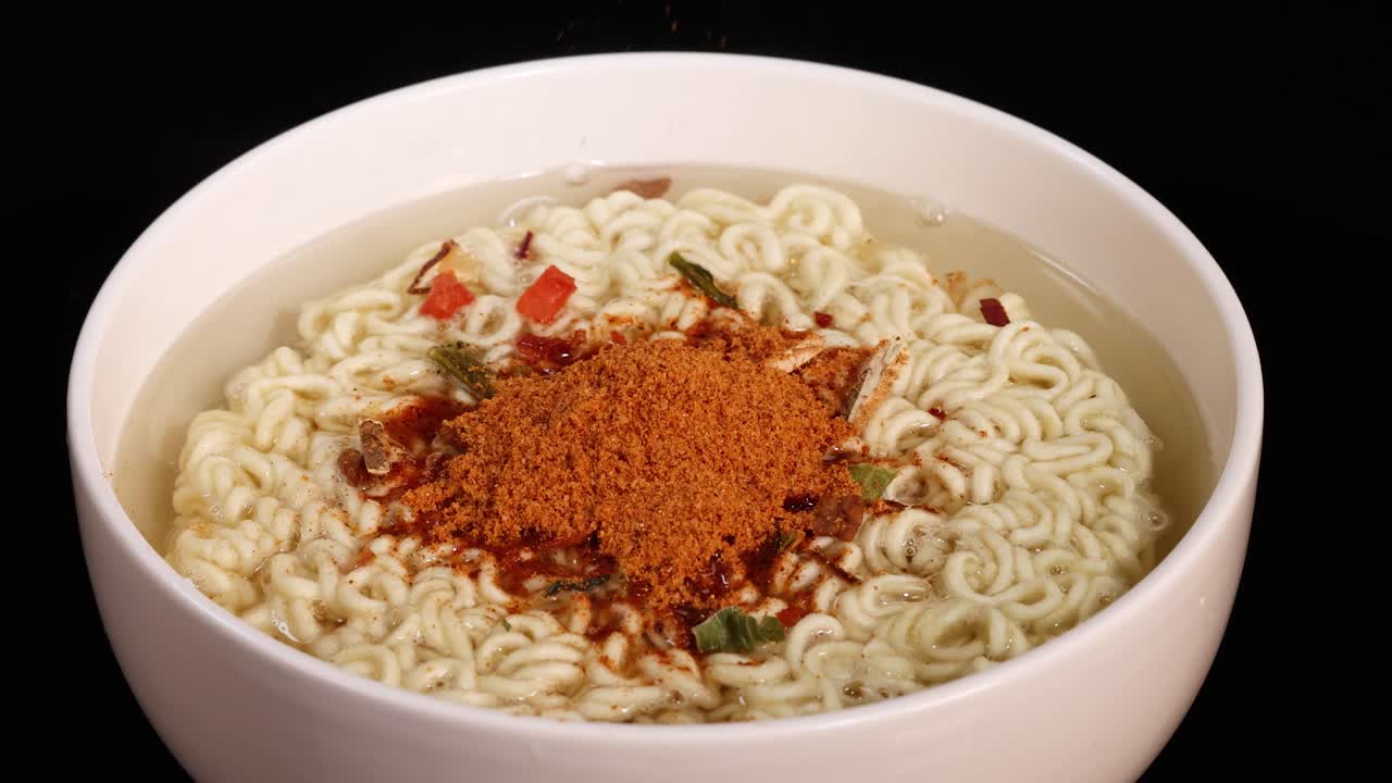 A close-up sequence shows dehydrated vegetables and spice powder being poured onto instant noodles in a white bowl, under bright studio lighting with a static camera