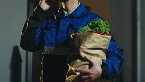 Primer-Plano-Del-Apuesto-Trabajador-Masculino-De-La-Entrega-Del-Supermercado-Hablando-Por-Teléfono-Y-Sosteniendo-Un-Paquete-Con-Verduras-Frescas