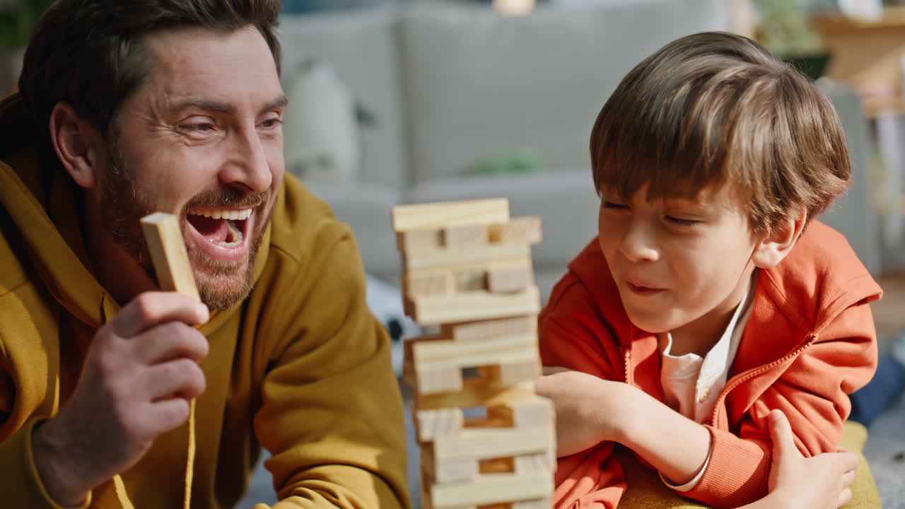 Primo piano: padre e figlio sorridenti che giocano a Jenga, sdraiati insieme sul pavimento.