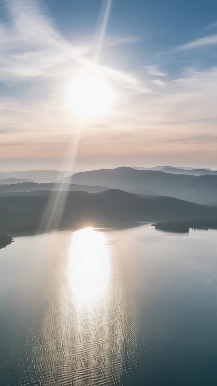 Video verticale: drone in planata che avanza, riprendendo il riflesso del sole all'alba sul lago con increspature e riverbero
