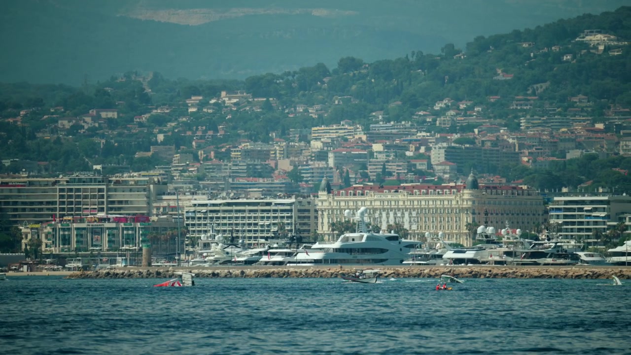 Lungomare di Cannes che mostra yacht di lusso ormeggiati nel porto con lo storico hotel Carlton e le colline di Antibes sullo sfondo, Cannes, Francia