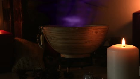 Detail-of-a-potion-making-witch-room,-with-purple-steam-coming-out-from-a-wooden-bowl,-with-animal-teeth-hanging-from-it,-next-to-candles-with-flickering-flames,-with-old-books-in-the-background