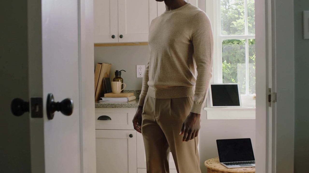 Open door revealing man in beige sweater shifting, glancing at laptop and tablet, preparing to work