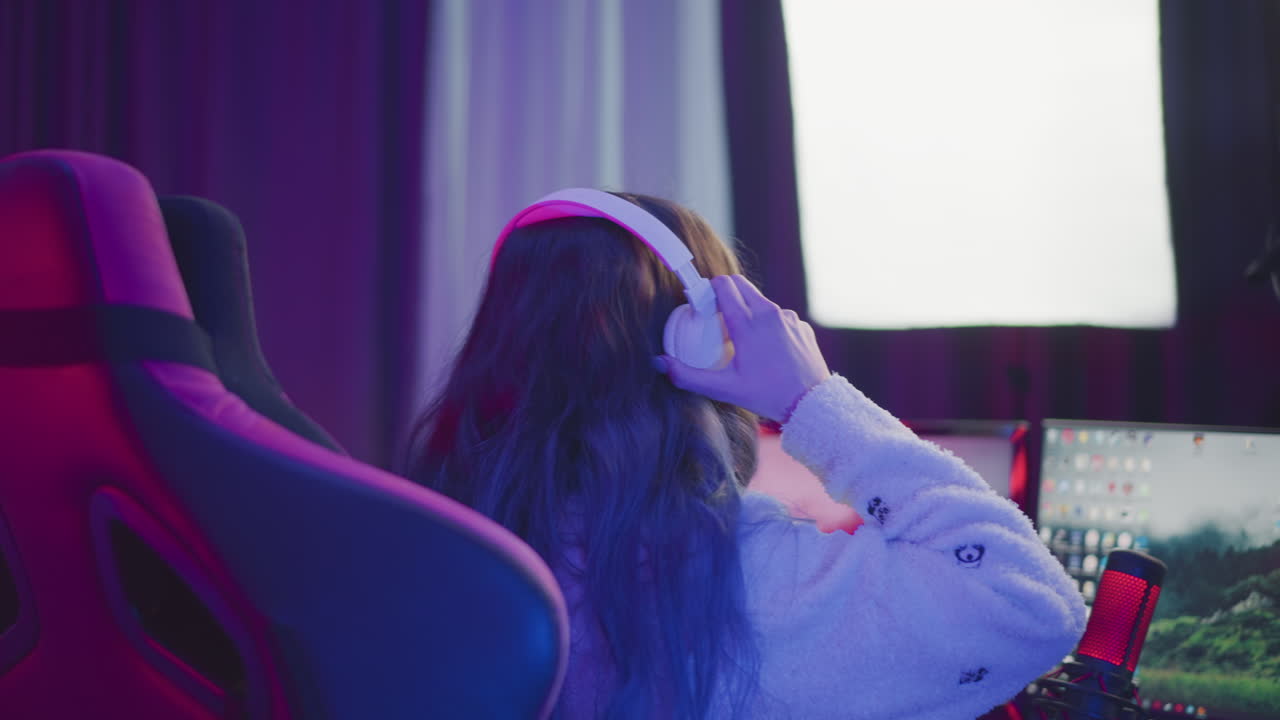 woman seated gaming chair with long hair wearing fluffy jacket reaches desk to pick up white headphone while red lit microphone and rgb keyboard illuminate area under moody light beside dual monitors