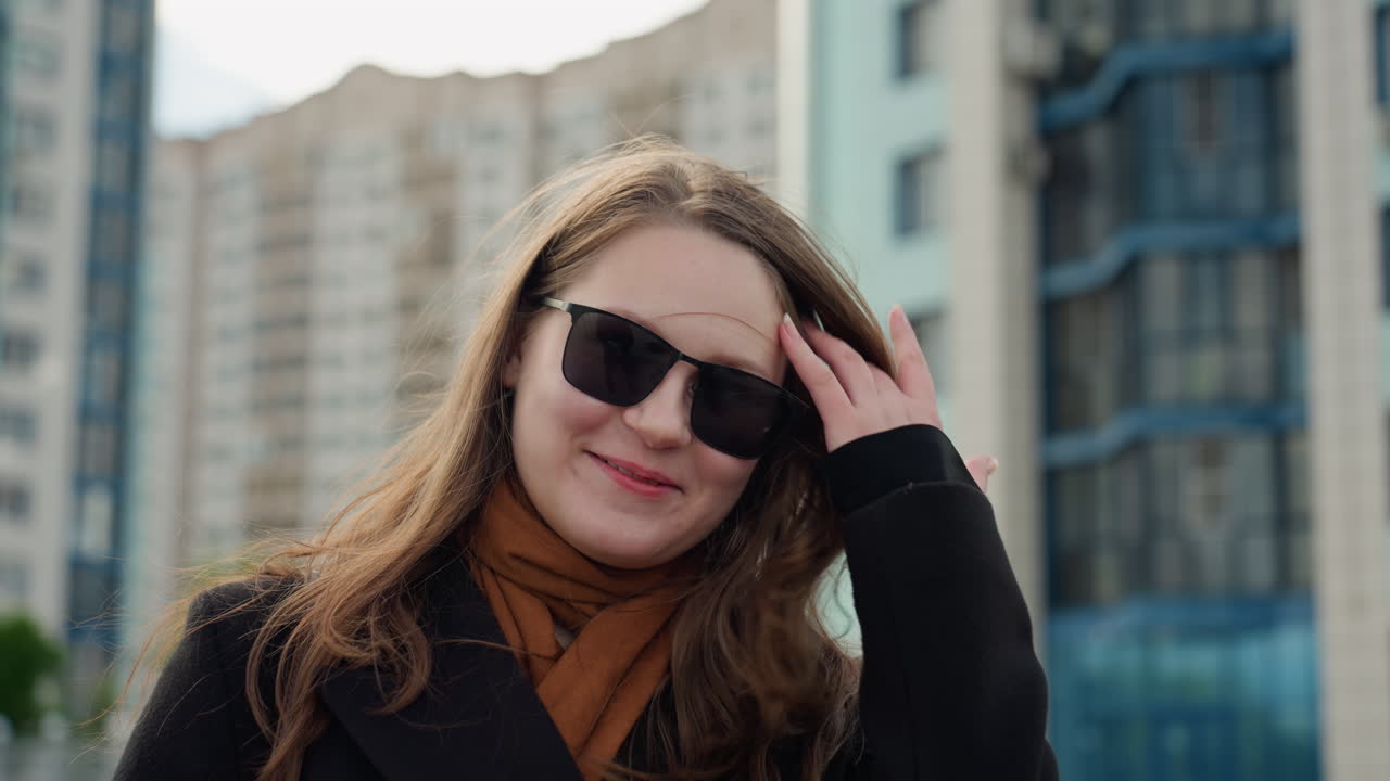 White lady wearing polarized sunglasses blushes, adjusting hair during outdoor portrait shoot in city, breeze lifts strands, warm scarf and coat visible, friendly vibe in blur modern buildings behind