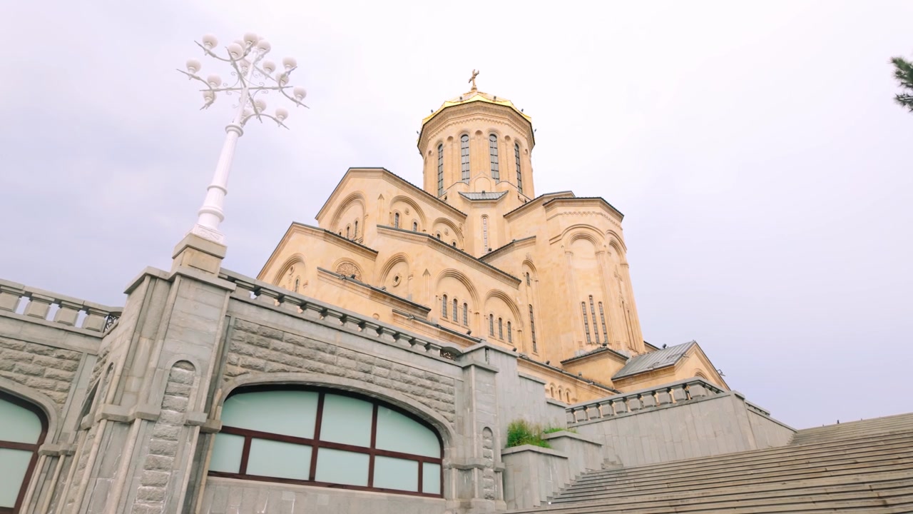 Vista impressionante para cima mostra a Catedral da Santíssima Trindade de Tbilisi, uma majestosa igreja ortodoxa georgiana com sua cúpula dourada característica e detalhes arquitetónicos intrincados contra um céu pálido.