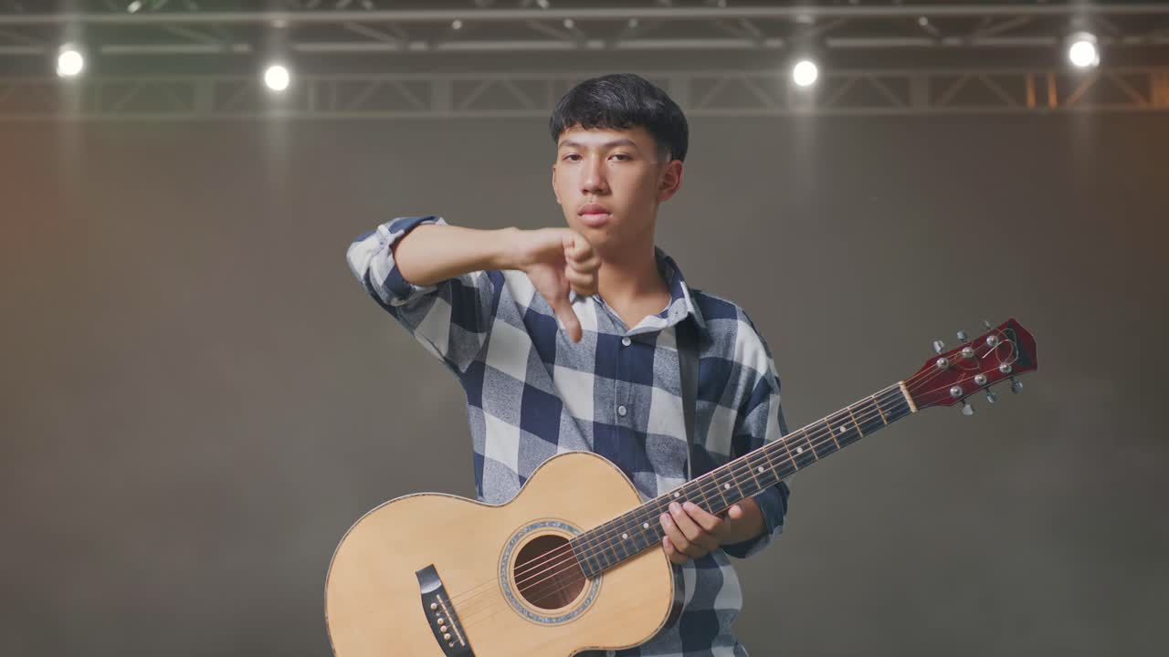 기타 를 들고 있는 아시아 청소년 이 무대 에 서 있는 동안 엄지손가락 을 내려 놓은 몸짓 을 하고 있다