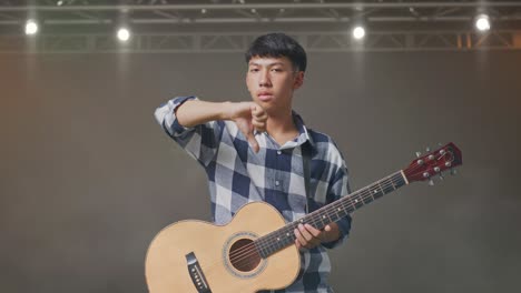 adolescente asiático con guitarra mostrando el pulgar hacia abajo mientras está de pie en el escenario