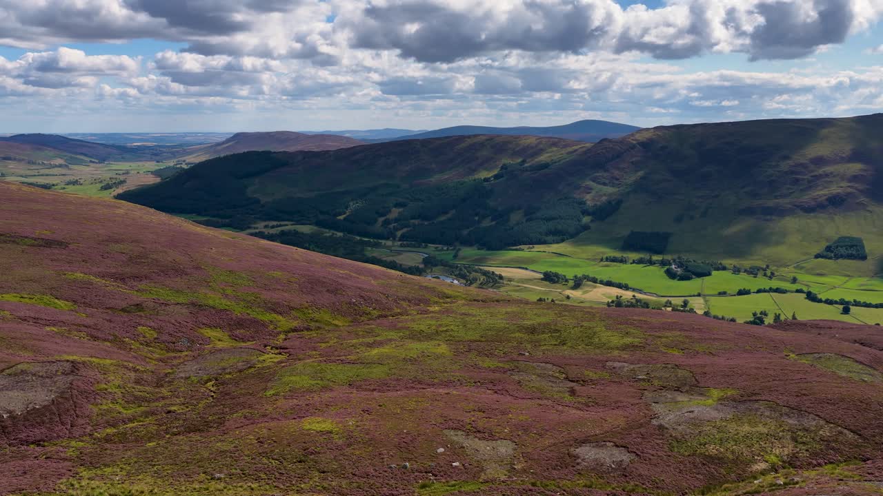 Drone camera smoothly pans across sunlit heather hills, green valley, and dramatic cloudy sky