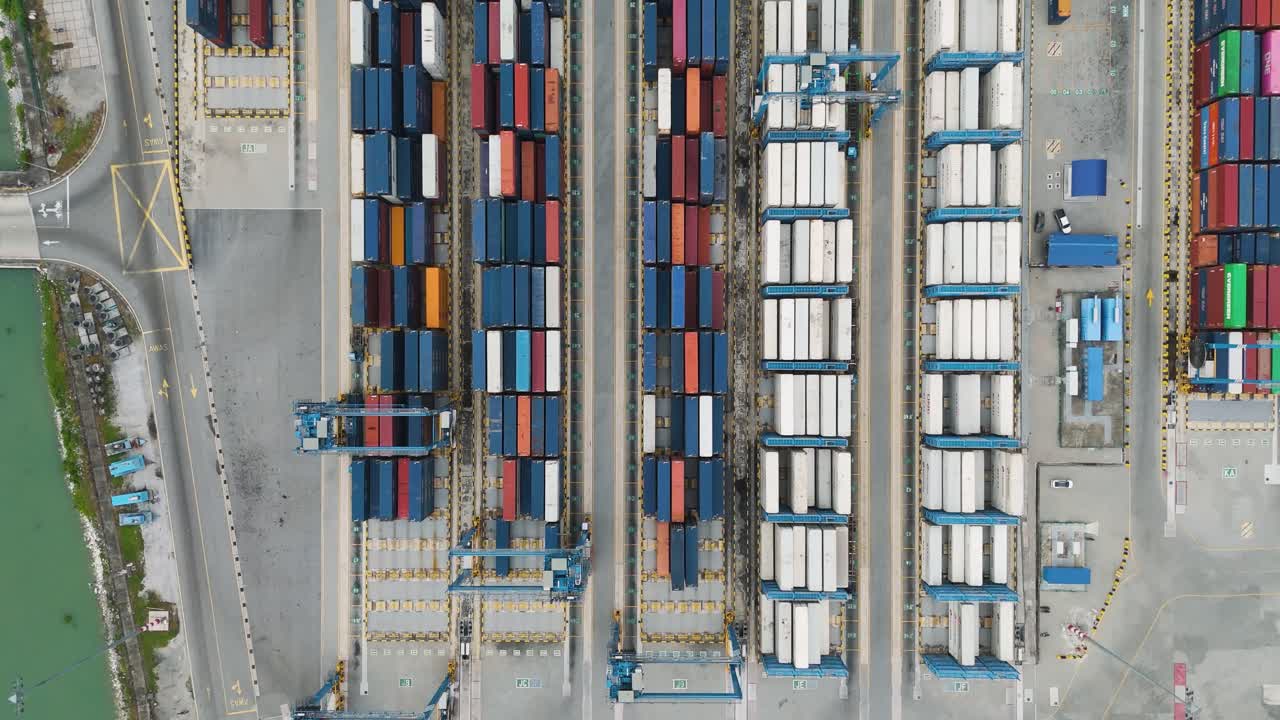 Top-down drone view of shipping containers and cargo trucks at a major Malaysian port, showing industrial logistics and organized freight transport operations in a container depot. UHD.