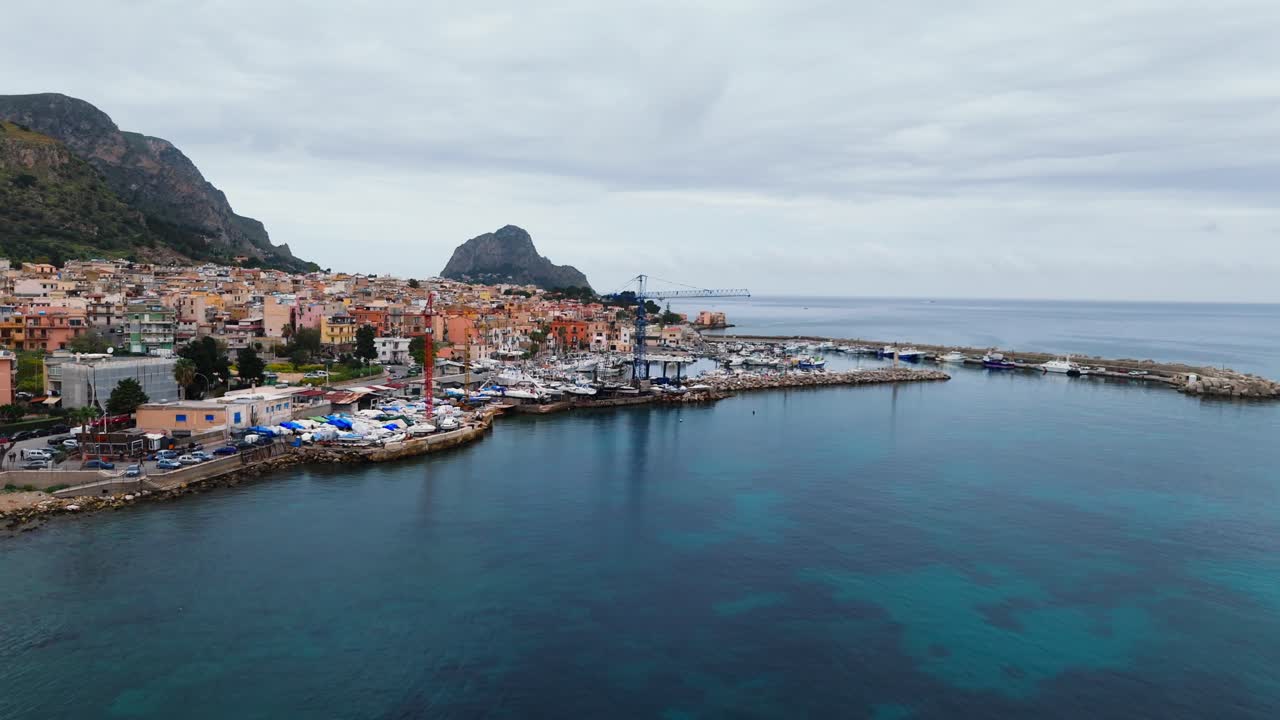 Coastal drone shot of Porticello, Sicily, featuring a harbor full of boats, colorful buildings, construction cranes and the Capo Zafferano rock formation in the distance