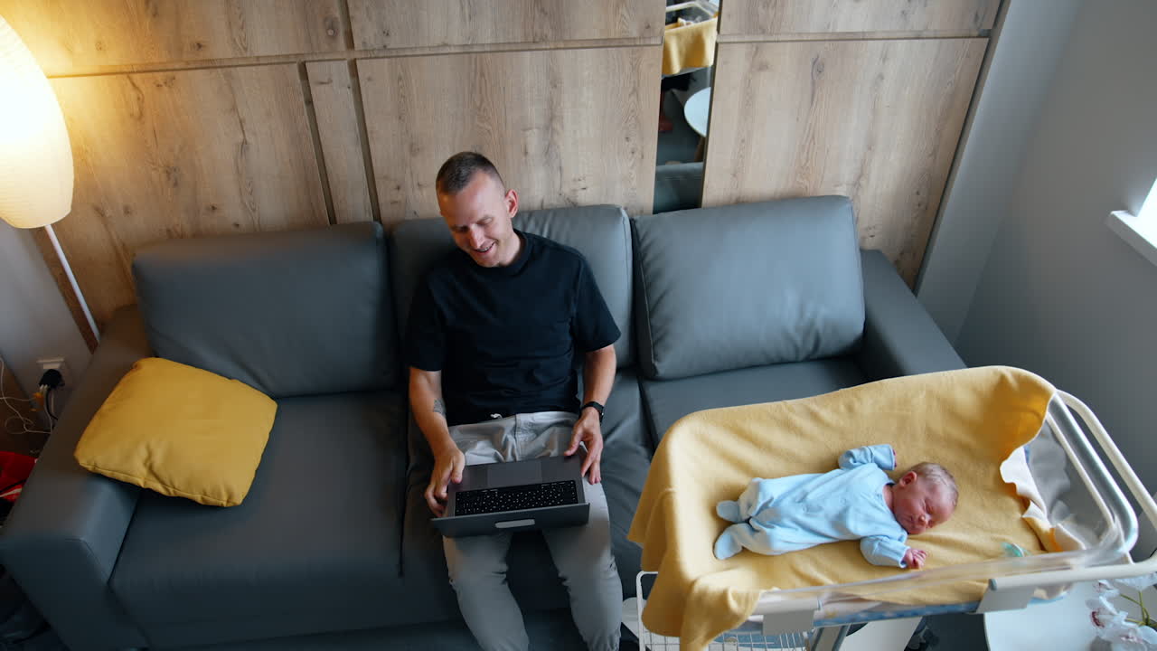 New-made father sitting on the sofa opens his laptop. Tiny newborn child lies in the crib beside. Top view.