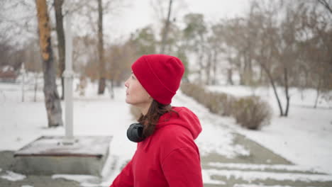 side view of athlete wearing red beanie and jacket walking through snowy park with headphones around neck and thoughtful expression, surrounded by bare trees, serene atmosphere, and winter scenery