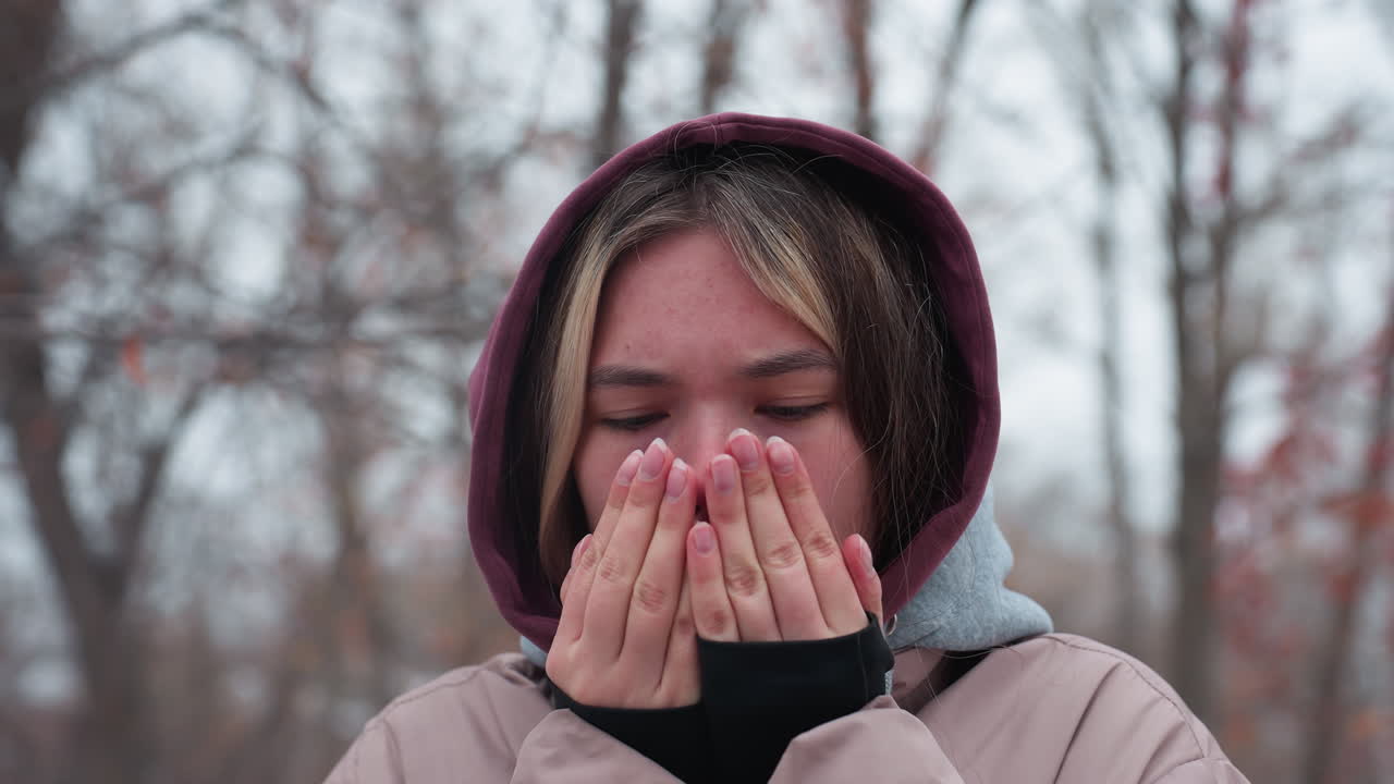 겨울 옷을 입은 여자가 추위에 서서 코를 문지르며 따뜻하게, 후드 재과 계층 옷은 추운 날씨를 강조합니다. 은 열매가 있는 나무의 흐릿한 배경.