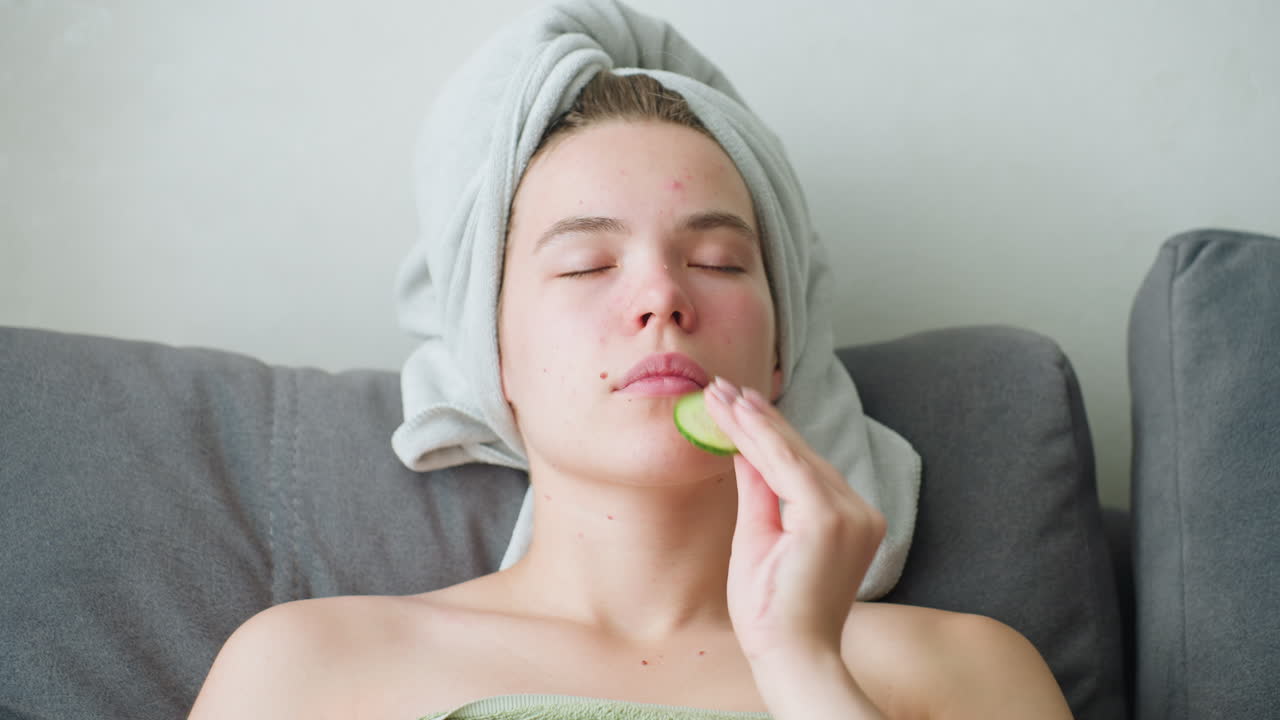 Close-up of woman with towel-wrapped hair gently pressing cucumber slice against cheek with closed eyes, engaging in calming skincare ritual under soft indoor light in peaceful, serene setting