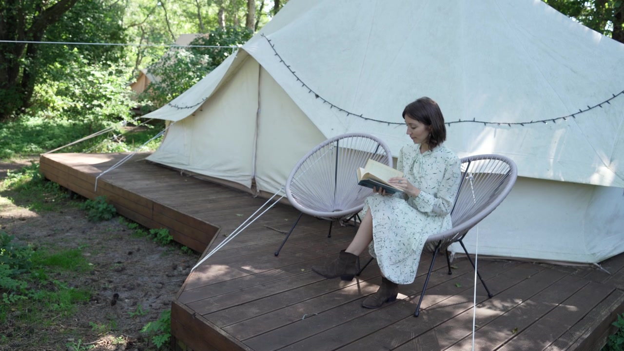 Ragazza con capelli scuri in abito campagnolo bianco legge un libro vicino a una tenda bianca in un glamping