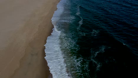 Vuelo-Aéreo-Suave-Sobre-El-Borde-Del-Agua-En-Una-Playa-De-Arena-Con-Olas-Rompientes