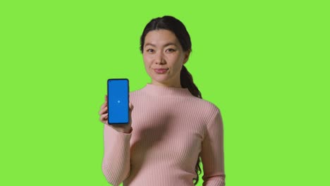 studio portrait of smiling woman holding blue screen mobile phone towards camera against green screen