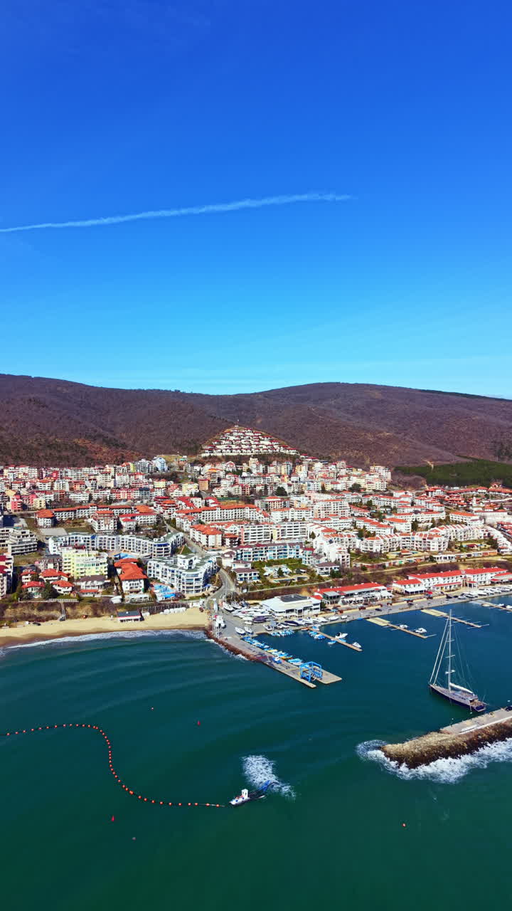 Vista costiera di una città sul mare con barche e cielo blu durante il giorno, che mostra edifici e colline sullo sfondo.