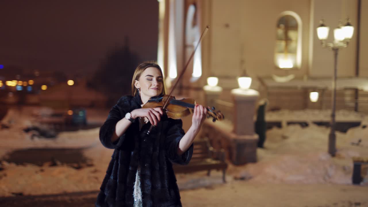 A beautiful girl is playing the violin in the centre of the city in the winter near the road on amid passing public transport. Blurred background.