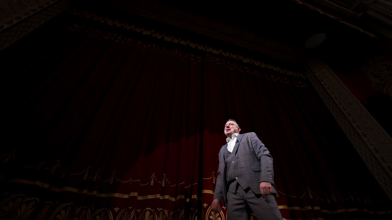 Actor performing on stage. Man in grey suit speaking with many gestures on red theatrical curtains background. View from below.