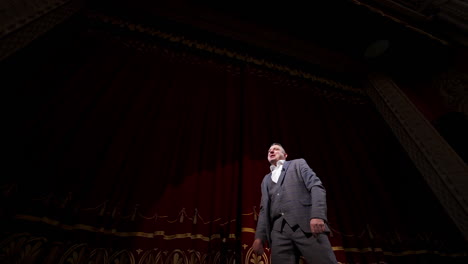 actor performing on stage. man in grey suit speaking with many gestures on red theatrical curtains background. view from below.