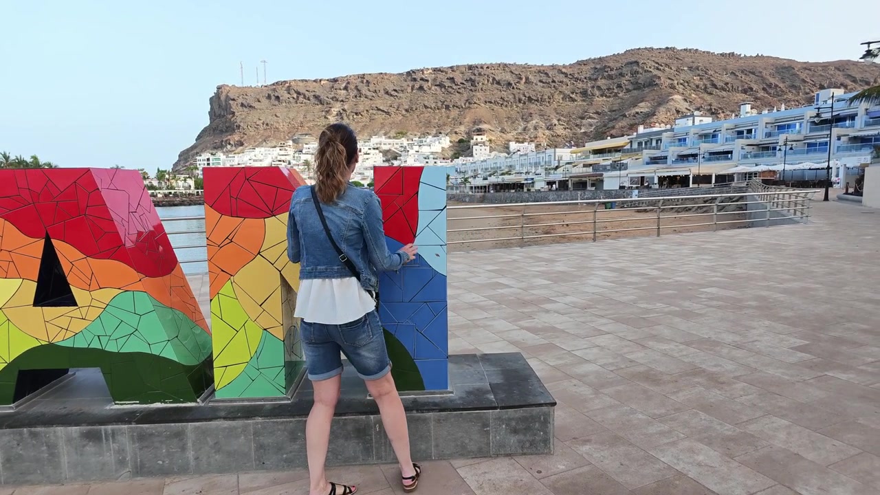 Donna turista che esplora il pittoresco porto di Puerto de Mogan. Cammina accanto al famoso cartello colorato con il nome della città.