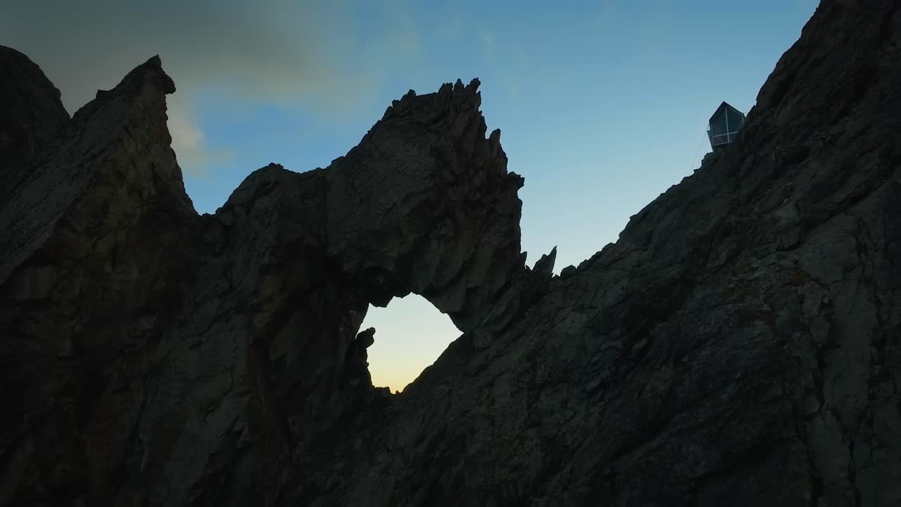 Aerial drone flying closer to a cliff in western Alps in Valle D'Asota during a sunny day with a hikers and climbers cabin standing on the steep cliff edge. The mountain tops are sharp and rocky.