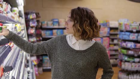 comida, conceito de saúde - mulher em um supermercado de pé em frente às prateleiras do congelador e escolhe comprar uma garrafa de iogurte, tomar um e colocá-lo no carrinho . câmera lenta. fechar