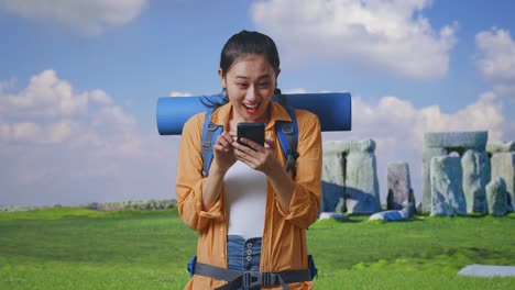 asian female hiker with mountaineering backpack looking at the smartphone and screaming goal celebrating the success while traveling in stonehenge