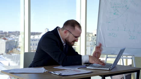 funny businessman typing on a laptop. office worker in glasses sitting at his desk in workplace and making mad face. problems in business.