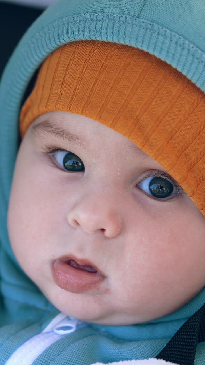 Adorable funny boy in warm clothes sits in a stroller. Sweet grey-eyed kid looks at camera calmly. Close up. Vertical video