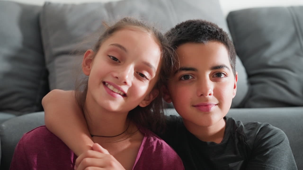 Precioso momento de hermanos sentados muy juntos en el sofá, sonriendo cálidamente con los brazos entrelazados, mostrando un fuerte vínculo y afecto en un acogedor ambiente hogareño con luz natural
