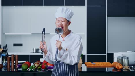 asian woman chef holding tongs and ladle and singing in home kitchen