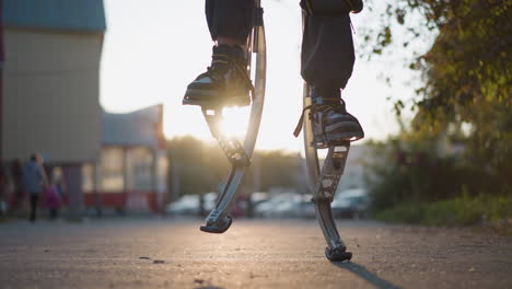 close-up view of legs on spring stilts walking in an outdoor urban setting with blurred parked cars, greenery, and shadows in the background. the person wears dark sneakers and pants