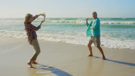 Vista-Lateral-De-Una-Mujer-Caucásica-Senior-Activa-Haciendo-Clic-En-Fotos-De-Un-Hombre-Senior-Con-Teléfono-Móvil-En-La-Playa