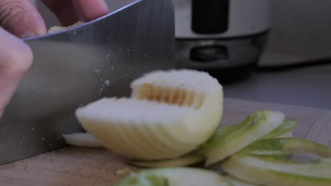 male hand holds skinned, cut and cored apple fruit with large metal knife on kitchen bench to cook stewed apple meal at home with skin off to side