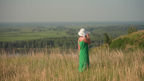 woman in white hat and green gown walks along grassy cliffside, turning to gaze across lush valley landscape with forest stretching into distant horizon under soft sunlight of clear sky