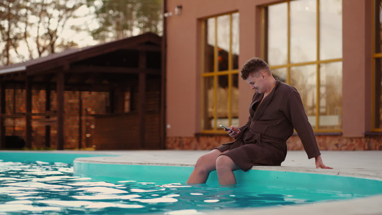 un joven despreocupado está descansando en un complejo de spa con piscina al aire libre navegando por internet por teléfono celular