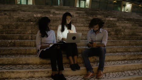 three young business people sitting on stairs with electronic devices in the evening on a windy day