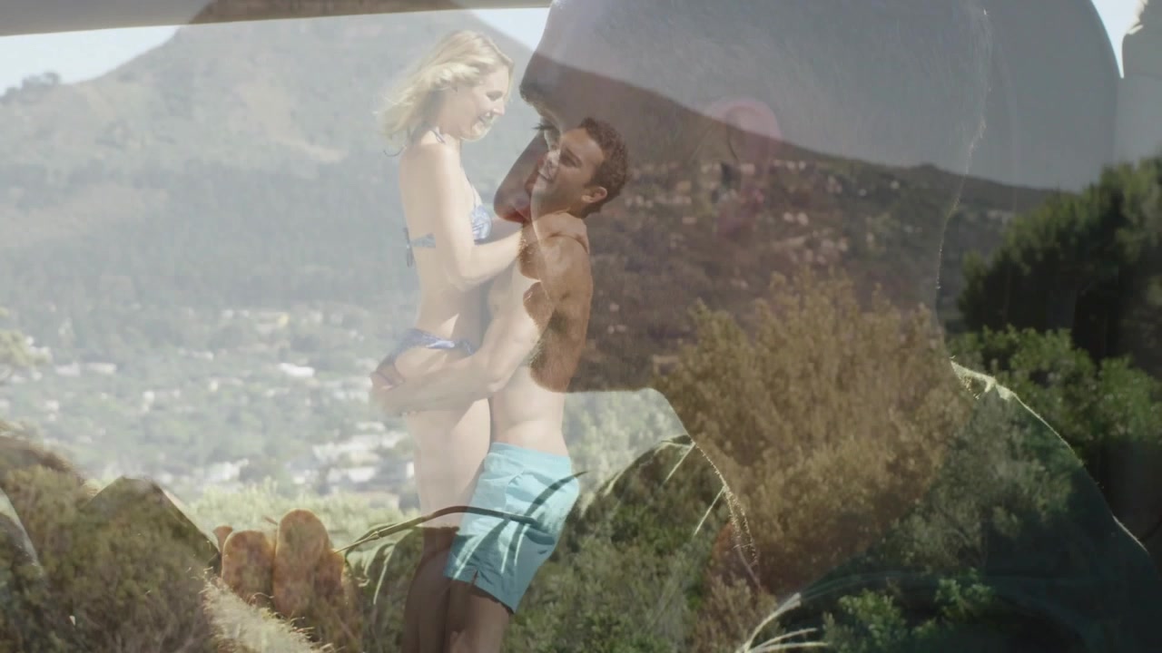 Uomo che solleva una donna in bikini su una collina soleggiata, la telecamera si sposta e una sovrapposizione di profilo collega una pubblicità di viaggio