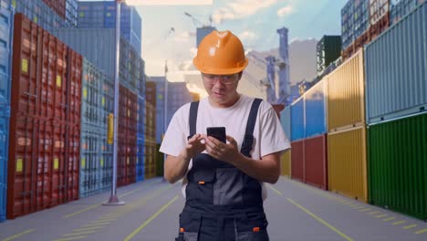 hombre asiático trabajador con gafas y casco de seguridad mirando su teléfono inteligente, sonriendo, y diciendo wow mientras estaba de pie en el almacén del patio de contenedores