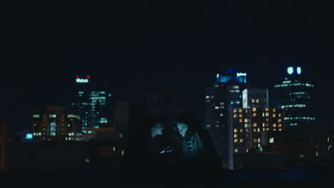 friends using smartphone mobile technology hanging out on rooftop at night enjoying weekend chatting sharing social media messages in urban city skyline background