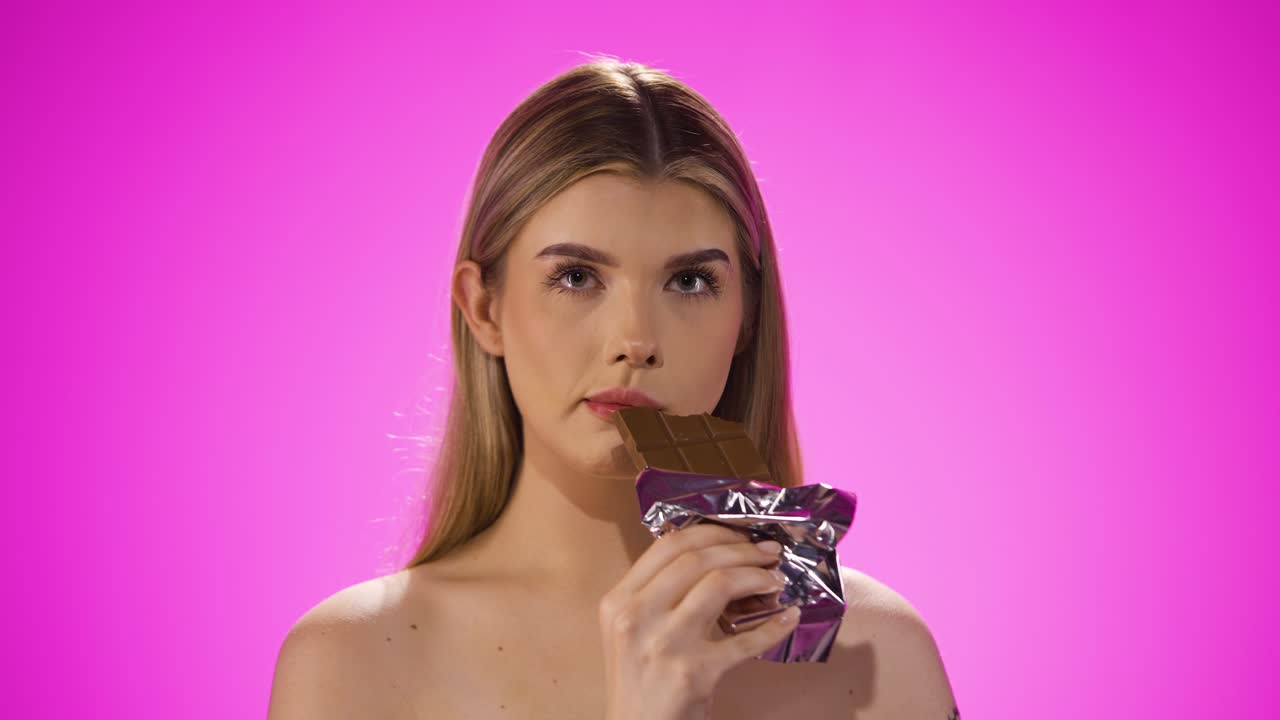 Medium shot of a beautiful young woman or model while she enjoys a bite of the vegan chocolate bar and loves different candy sweets against purple background in slow motion