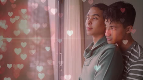 Animation-of-glittering-hearts-over-happy-diverse-gay-male-couple-looking-out-of-window,-embracing